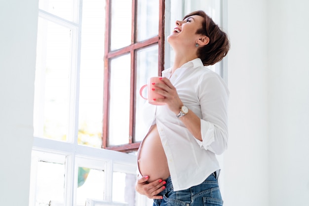 Heureuse femme enceinte tient son ventre et rit près de la fenêtre ouverte