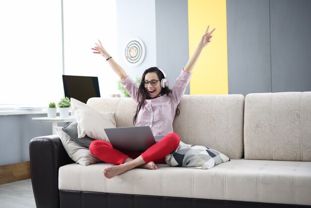 Heureuse femme est assise sur un canapé portant des écouteurs levant joyeusement ses mains