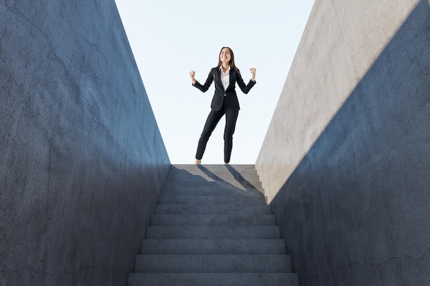 Heureuse femme européenne célébrant le succès au sommet d'escaliers en béton abstraits avec la lumière du soleil.