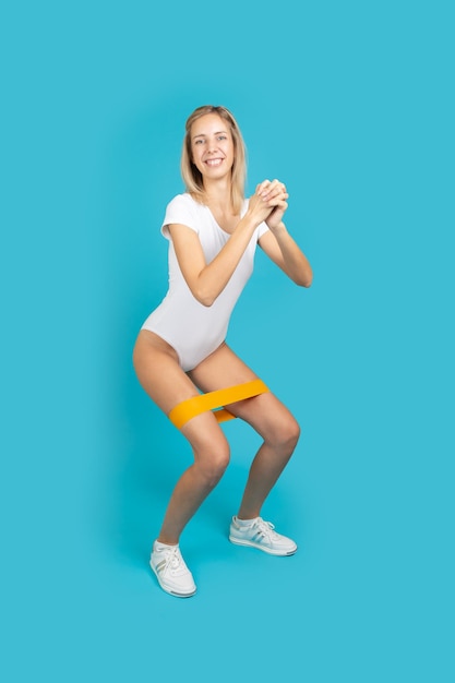 Heureuse femme faisant des exercices de squat ayant une bande de résistance orange sur les jambes en body blanc sur fond turquoise Faire du sport pour être en forme Améliorer sa santé par soi-même à la maison