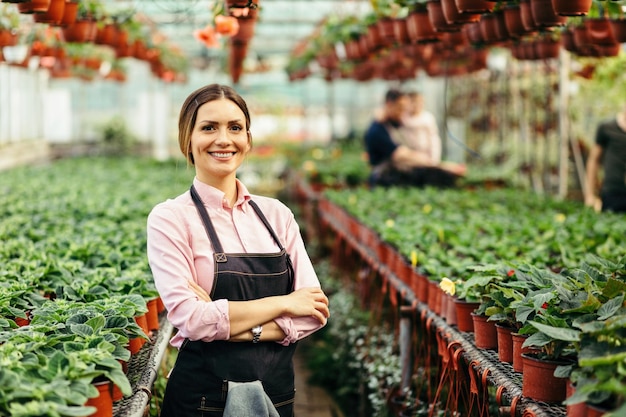 Heureuse femme fleuriste debout avec les bras croisés et regardant la caméra à la pépinière Il y a des gens en arrière-plan