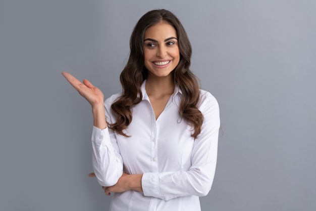 Heureuse femme gestionnaire professionnelle en tenues de soirée montre la main ouverte pour l'arrière-plan gris de l'espace de copie, présentant le produit.