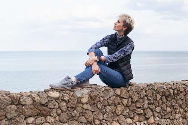 Heureuse femme hipster assise sur le trottoir de pierre près de la mer en automne