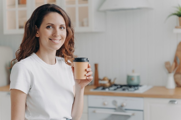 Heureuse femme hispanique tenant une tasse de café jetable souriant regardant la caméra dans une cuisine moderne