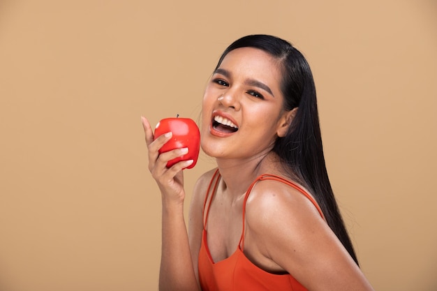 Heureuse femme indienne asiatique avec de longs cheveux raides noirs et un grand sourire de dent tenant Apple à la main et un panier avec des fruits frais sains et des dents blanches souriantes. Espace de copie de fond jaune beige