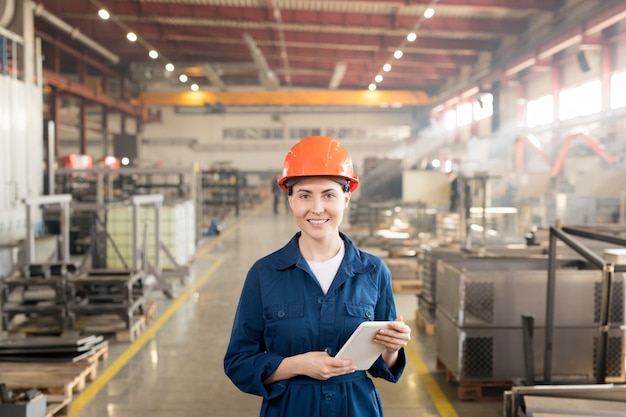 Heureuse femme ingénieur en casque et salopette vous regardant tout en travaillant à l'intérieur d'une grande usine industrielle
