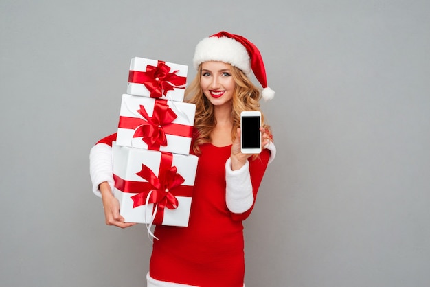 Heureuse femme joyeuse en tenue de père noël rouge montrant un téléphone portable à écran blanc isolé sur la surface grise