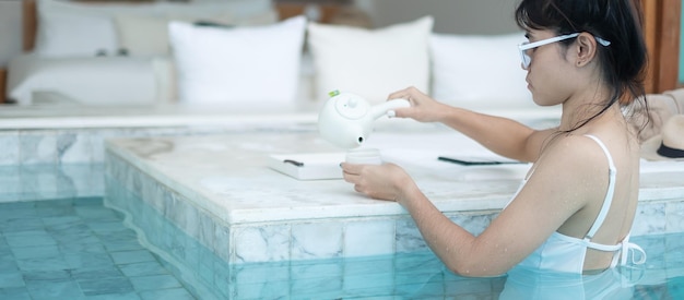 Heureuse femme en maillot de bain blanc buvant du thé chaud dans un hôtel de luxe avec piscine jeune femme avec chapeau profiter dans un complexe tropical Détente été voyage vacances vacances et week-end concept