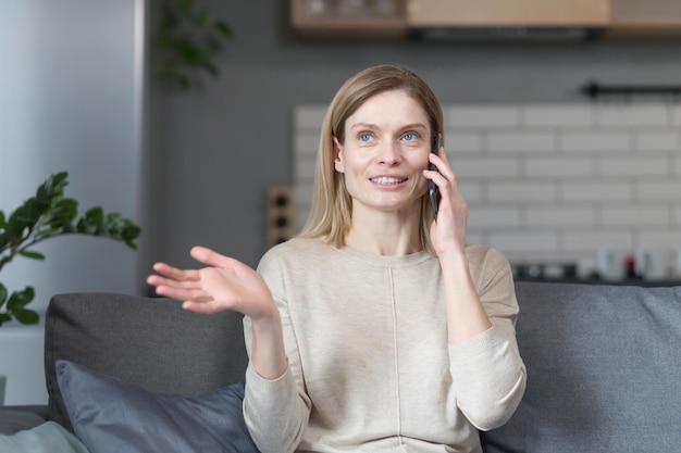 Heureuse femme à la maison assise sur le canapé et parlant au téléphone