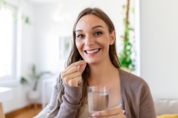 Heureuse femme à la maison assise à table à la maison tenant une tasse d'eau et de comprimés traitement du coronavirus gros plan arrière-plan flou