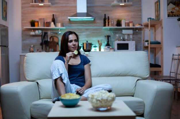 Heureuse femme mangeant du pop-corn sur un canapé et regardant la télévision dans le salon à la maison. Dame seule excitée, amusée, profitant de la soirée assise sur un canapé confortable vêtue d'un pyjama devant la télévision.