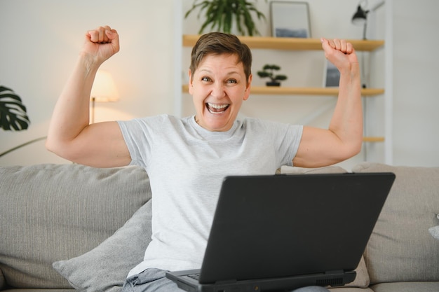 Heureuse femme mature aux cheveux gris excitée célébrant la victoire en ligne à l'aide d'un ordinateur portable regardant l'écran assis sur le canapé à la maison femme d'âge moyen se sentant étonnée surprise par une incroyable bonne nouvelle