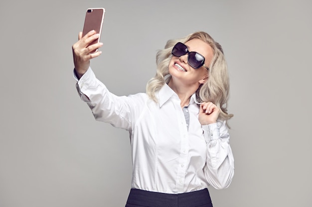 Heureuse femme mature à lunettes de soleil prend un selfie à partager sur les réseaux sociaux sur gris isolé