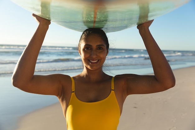 Heureuse femme métisse sur la plage portant une planche de surf sur la tête. loisirs en plein air sains au bord de la mer.