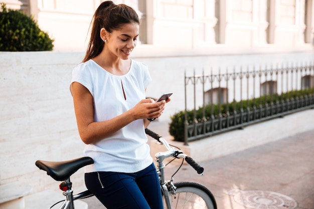 Heureuse femme mignonne tapant mesaage sur le smartphone s'appuyant sur un vélo dans la rue