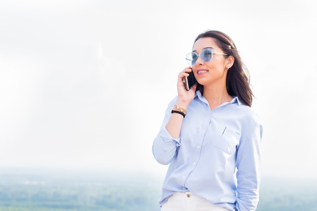 Heureuse femme à la mode avec des lunettes parlant au téléphone et souriant au fond de la nature