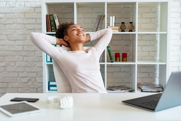 Heureuse femme mulâtre ayant une pause au bureau, les bras croisés par la tête et allongée sur la chaise en levant les yeux