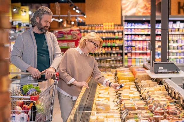 Heureuse femme mûre prenant morceau de fromage emballé à partir de l'affichage tout en marchant le long des produits laitiers avec son mari en supermarché