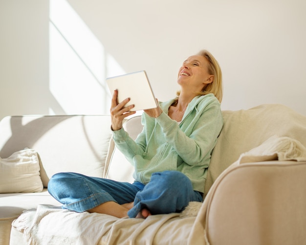 Heureuse femme mûre profitant du temps à la maison tout en se relaxant sur un canapé et en regardant une vidéo en numérique