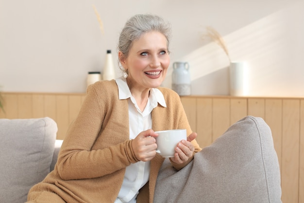 Heureuse femme mûre reposant sur un canapé confortable, boire du café ou du thé, regarder ailleurs, se détendre sur un canapé confortable à la maison, savourer une boisson chaude