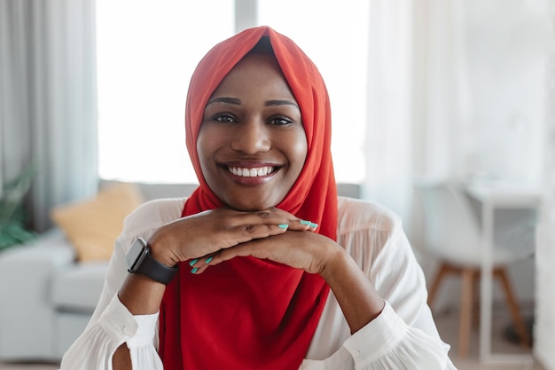 Photo heureuse femme musulmane afro-américaine en hijab rouge assise sur le lieu de travail au bureau à domicile regardant la caméra et souriant