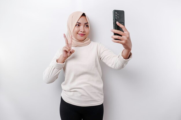 Une heureuse femme musulmane asiatique portant un foulard tenant son téléphone isolé par fond blanc