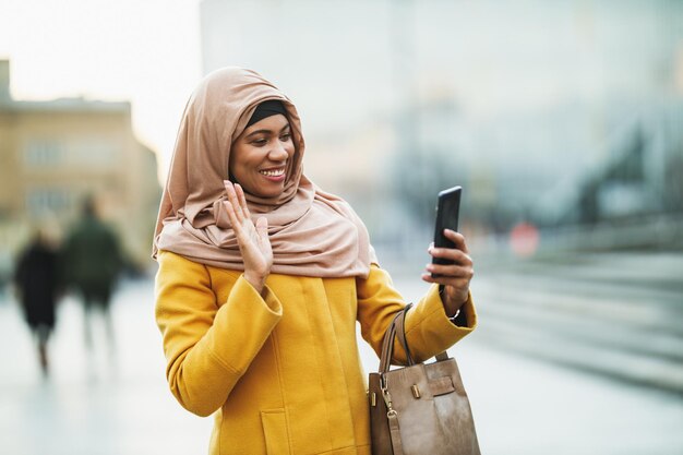 Heureuse femme musulmane noire portant un hijab et utilisant son smartphone en marchant dans le centre-ville.