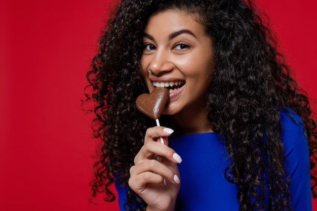 Heureuse femme noire avec des bonbons au chocolat en forme de coeur souriant sur mur rouge