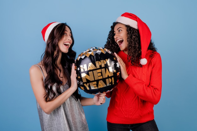 Heureuse femme noire et dame caucasienne avec ballon de bonne année et chapeaux de Noël isolés sur bleu