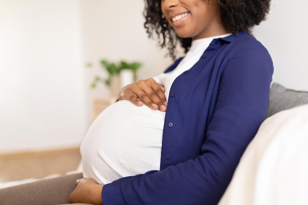 Heureuse femme noire millénaire bouclée en vêtements domestiques avec un gros ventre aime la grossesse est assise sur le lit