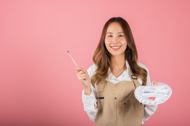 Heureuse femme peignant à l'aide d'un pinceau et d'une palette de couleurs, belle jeune femme artiste asiatique tenant un pinceau et une palette de peinture, prise de vue en studio isolée sur fond rose, Peintures et équipement d'art