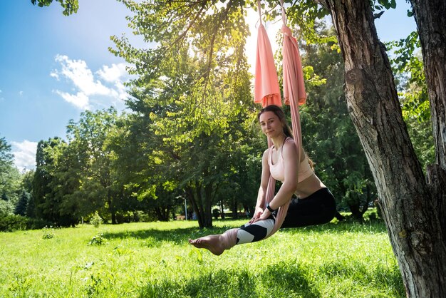 Heureuse femme physiquement développée pratique le yoga sur des toiles aériennes sur un arbre