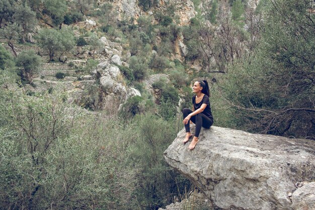 Heureuse femme pieds nus en vêtements noirs souriant et observant la chaîne de montagnes tout en étant assise sur un rocher dans la vallée de Majorque, Espagne