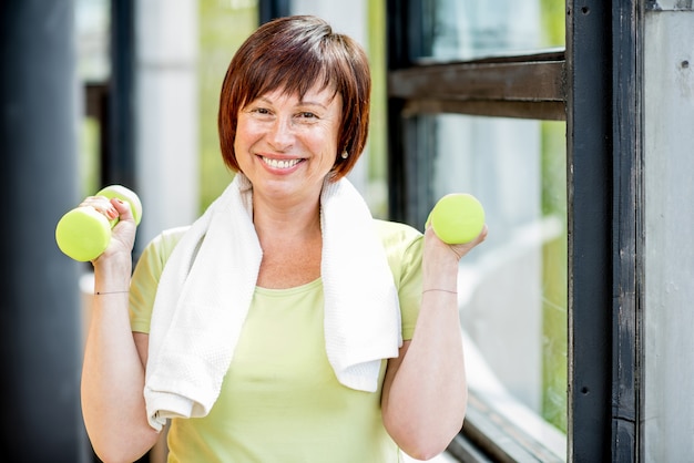 Heureuse Femme Plus âgée Dans La Formation De Vêtements De Sport Avec Des Haltères à L'intérieur Sur Le Fond De La Fenêtre