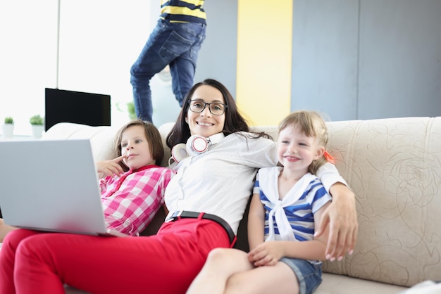 Heureuse femme posant avec ses enfants sur un canapé tout en travaillant sur un ordinateur portable