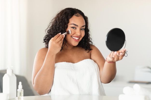 Heureuse femme positive au corps noir avec des cheveux bouclés dans une serviette regardant dans le miroir et appliquant de la poudre avec un pinceau sur le visage