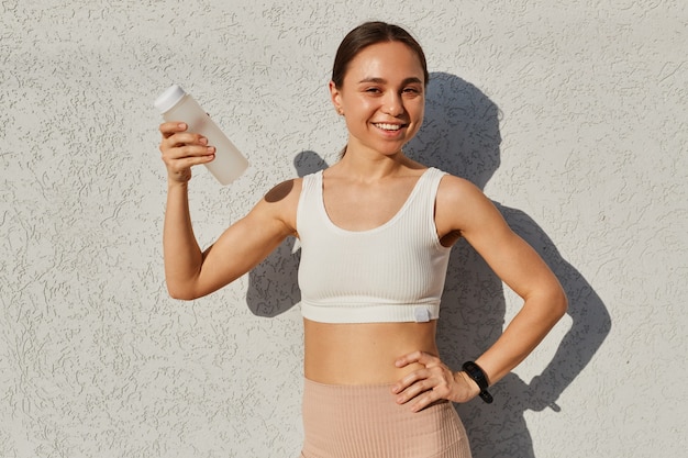 Heureuse femme positive portant un haut sportif blanc aux cheveux noirs, tenant une bouteille d'eau à la main, regardant la caméra en souriant, a soif après l'entraînement.