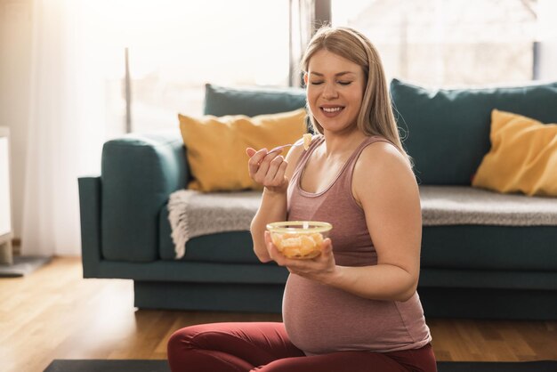 Heureuse femme qui attend de manger une salade de fruits frais tout en faisant de l'exercice à la maison. Elle se détend sur un tapis d'exercice dans son salon le matin.