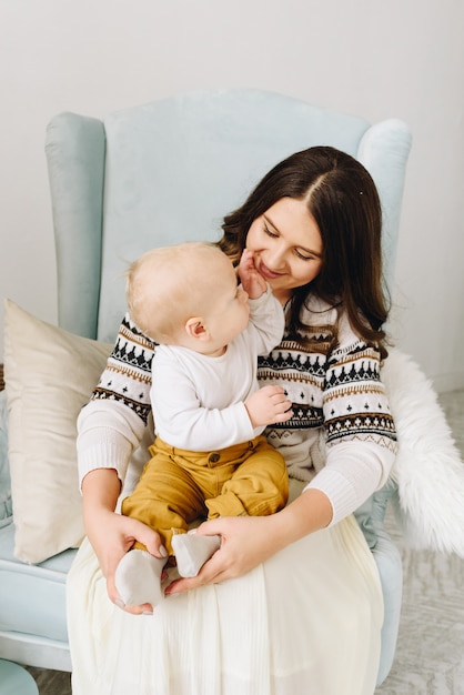 Heureuse femme de race blanche assise dans un fauteuil et tenant son bébé, portant un pull de Noël festif