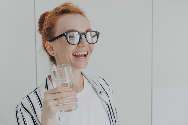 Heureuse femme riant au gingembre debout dans la cuisine le matin avec un verre d'eau minérale pure