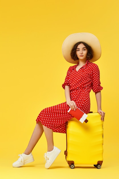 Heureuse femme en robe rouge avec passeport assis sur une valise sur fond jaune.