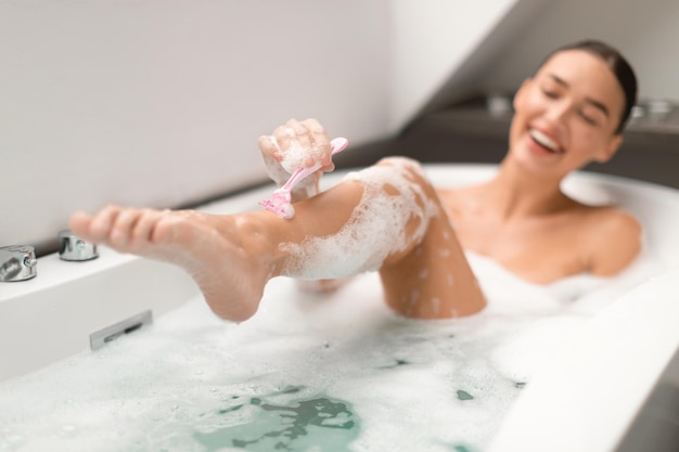 Heureuse femme se rasant les jambes se baignant dans la salle de bain moderne Mise au point sélective