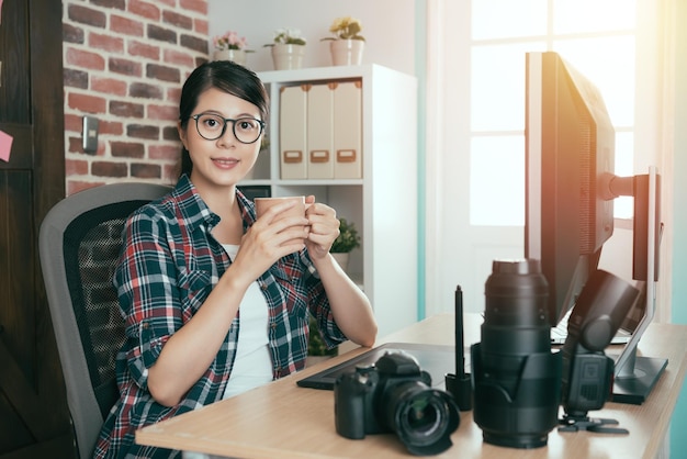 heureuse femme séduisante photographe face à la caméra souriante et assise sur le bureau travaillant en buvant du café expresso chaud se reposant à l'heure du thé de l'après-midi.