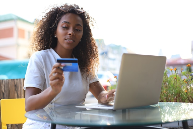 Photo heureuse femme séduisante utilisant une carte de crédit et un ordinateur portable dans un café en plein air.