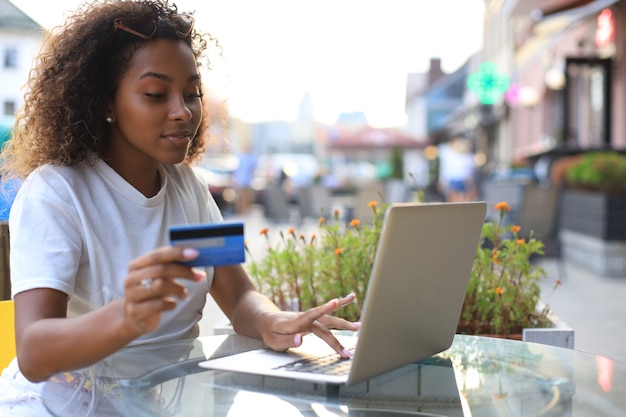 Photo heureuse femme séduisante utilisant une carte de crédit et un ordinateur portable dans un café en plein air.