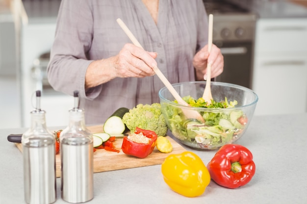 Heureuse femme senior jetant une salade debout dans la cuisine