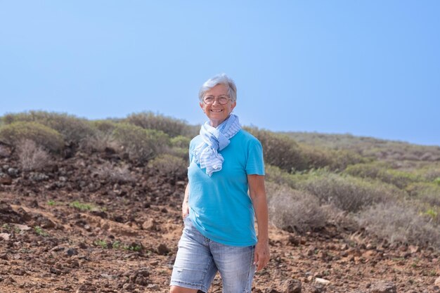 Heureuse femme senior mature marchant en plein air dans la campagne portant des lunettes et une écharpe bleue