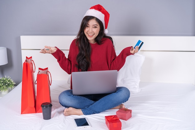 Heureuse femme shopping en ligne pour cadeau de Noël avec ordinateur portable et carte de crédit sur lit