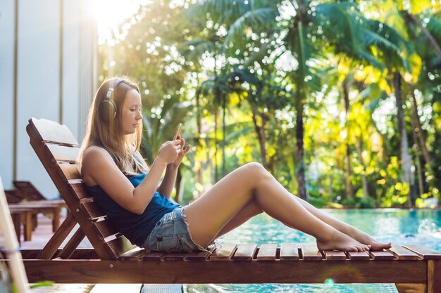 Heureuse femme smartphone se relaxant près de la piscine, écoutant avec des écouteurs de la musique en streaming. Belle fille utilisant les données 4g de son application pour téléphone portable pour jouer des chansons tout en se relaxant pendant les vacances d'été de luxe