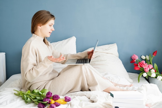 Heureuse femme souriante assise sur le lit en pyjama, avec plaisir en appréciant les fleurs, en discutant à l'aide d'un ordinateur portable